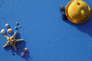 Frame with beach accessories on a nautical theme yellow straw hat, sunglasses, starfish and shells on a blue background. Vacation concept, sea trip, UV protection, swimming. Copy space. Flatlay photo