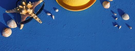 Frame with beach accessories on a nautical theme yellow straw hat, sunglasses, starfish and shells on a blue background. Vacation concept, sea trip, UV protection, swimming. Copy space. Flatlay photo