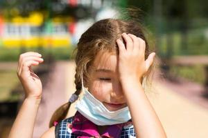 Girl with a backpack near the school after classes takes off a medical mask, unhappy, tired and headache. He holds his head with his hands, there is shortage of oxygen. Coronavirus, security measures photo