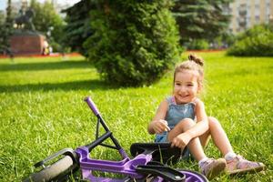 Cheerful girl sitting resting on the grass in a park near to the purple running bike in summer time. Children's active entertainment, scooter for toddlers, happy child. Copy space. Summer background photo