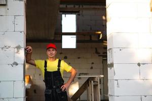 Construction worker engineer at construction site measures the length of window opening and brick wall with tape measure. Man in work clothes - jumpsuit and cap photo