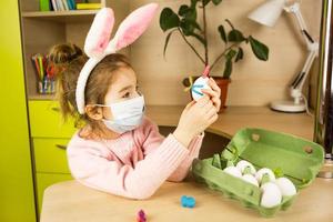 A girl in the ears of a hare makes an Easter bunny in a medical mask out of an egg and plasticine. DIY sitting at home, preparing for a religious holiday, home interior.