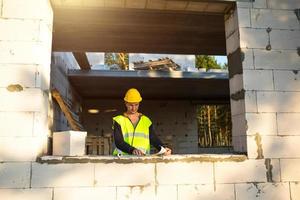 A construction engineer and designer in a yellow hardhat studies a drawing of a building on a construction site. A house made of porous concrete blocks, a worker in the process at the window and wall photo