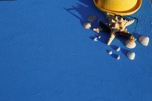 Frame with beach accessories on a nautical theme yellow straw hat, sunglasses, starfish and shells on a blue background. Vacation concept, sea trip, UV protection, swimming. Copy space. Flatlay photo