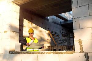A construction engineer and designer in a yellow hardhat studies a drawing of a building on a construction site. A house made of porous concrete blocks, a worker in the process at the window and wall photo