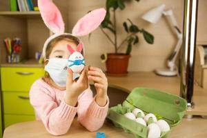 A girl in the ears of a hare makes an Easter bunny in a medical mask out of an egg and plasticine. DIY sitting at home, preparing for a religious holiday, home interior. photo