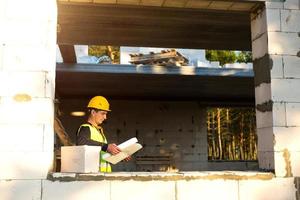 A construction engineer and designer in a yellow hardhat studies a drawing of a building on a construction site. A house made of porous concrete blocks, a worker in the process at the window and wall photo