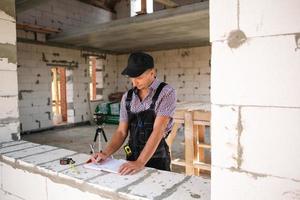 Construction worker engineer at construction site measures the length of window opening and brick wall with tape measure. Man in work clothes - jumpsuit and cap photo