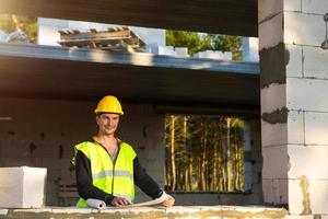 A construction engineer and designer in a yellow hardhat studies a drawing of a building on a construction site. A house made of porous concrete blocks, a worker in the process at the window and wall photo