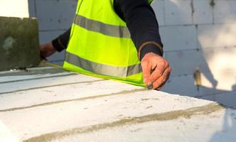 Construction worker at construction site measures the length of the window opening and the wall with tape measure. Cottage are made of porous concrete blocks, protective clothing - hardhat and a vest photo