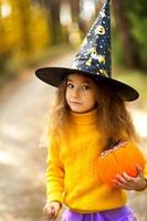 A girl in a witch's hat, in a yellow sweater and with a pumpkin with carved eyes and a mouth Jack o Lantern in a yellow autumn forest. Halloween Holiday photo