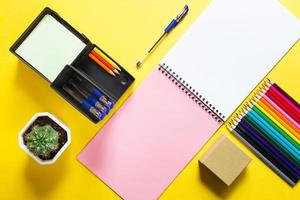 Layout of multi-colored stationery on a yellow background spiral notebook, colored pencils, stand, pens, indoor flower. Business flat lay, mock up. Sheets for notes, office work. Back to school photo