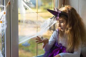 una niña disfrazada de bruja en el alféizar de la ventana está jugando con una telaraña y una araña, decorando la casa para la fiesta de halloween. terrible paisaje, miedo y horror, calabazas