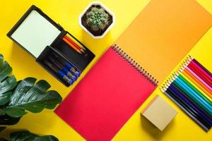 Layout of multi-colored stationery on a yellow background spiral notebook, colored pencils, stand, pens, indoor flower. Business flat lay, mock up. Sheets for notes, office work. Back to school