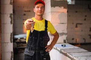 Construction and finishing worker holds out key to the house. Turnkey construction, repair, relocation, mortgage. Engineer in protective workwear jumpsuit and cap photo