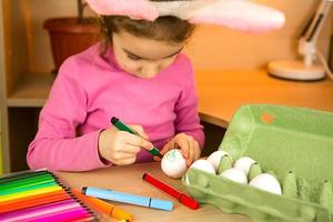 A girl in the ears of an Easter bunny paints eggs with a felt-tip pen in the home interior. Crafts, preparation for a religious holiday, a tray with eggs, hare ears made of plasticine photo