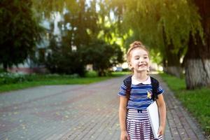A little girl of Caucasian appearance in a school uniform with a backpack and the book and a set of markers. back to school. Elementary school, developing activities for preschoolers. Space for text photo