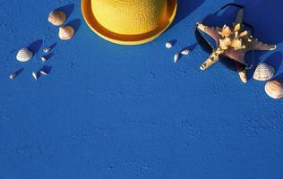 Frame with beach accessories on a nautical theme yellow straw hat, sunglasses, starfish and shells on a blue background. Vacation concept, sea trip, UV protection, swimming. Copy space. Flatlay photo