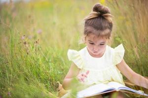 una chica con un vestido amarillo se sienta en la hierba sobre una manta en un campo y lee un libro de papel. día internacional del niño. horario de verano, infancia, educación y entretenimiento, núcleo rural. copie el espacio foto