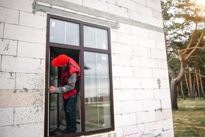 el instalador de ventanas ajusta las bisagras y revisa las nuevas ventanas en la cabaña en construcción. ordenar ventanas con disposición dorada y laminación marrón para una casa particular. un trabajador en uniforme foto