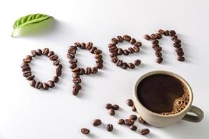 taza de café y granos de café sobre fondo blanco con texto abierto hecho de granos de café foto