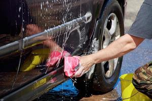 el hombre lava la rueda del coche. la mano masculina sostiene una esponja rosa con espuma jabonosa para limpiar. foto