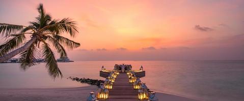 increíble cena romántica en la playa en una terraza de madera con velas bajo el cielo del atardecer. romance y amor, cenas de destino de lujo, mesa exótica con vista al mar foto