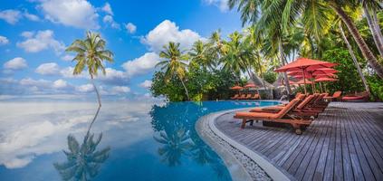 Panoramic holiday landscape. Luxurious beach resort hotel swimming pool and beach chairs or loungers under umbrellas with palm trees, blue sunny sky. Summer island seaside, travel vacation background photo