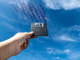Hand holding a Floppy A disc, insert the reader. to read and write data It is an old technology with a square shape. the background is the sky There is digital information flowing out. photo