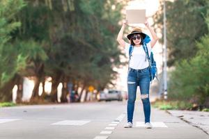 sonrisa feliz autostopista mujer asiática mochila esperando el transporte en automóvil durante el fin de semana para viajar y viajar en vacaciones al atardecer foto