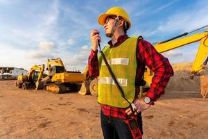 ingeniero civil técnico asiático habla con el equipo por walkie talkie y señala el punto de inspección para el proyecto de trabajo de construcción de transporte con excavadora en el fondo del sitio de ingeniería foto