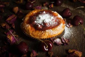 Cherry jam tart with raw cherries laid around and dried roses on wooden boards on black background. photo