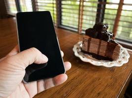 pastel de chocolate con la mano de un joven hombre de negocios sosteniendo un teléfono con una pantalla en blanco para publicidad, ingrese un mensaje o un tema de promoción en la cafetería. foto