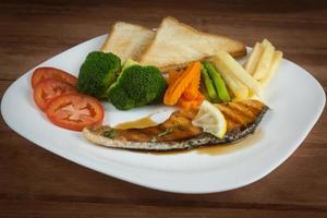 Grilled salmon steak slices until cooked, placed on a white plate with lemon on fish and vegetables, placed around the plate on a white wooden floor. photo