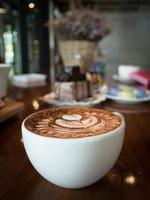 capuchino con espuma de leche fresca y arte latte en un vaso de cerámica blanca sobre una mesa de madera en una cafetería. foto