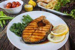 bistec de salmón con salsa en un plato blanco con limón en el plato y papas fritas crujientes. muchas verduras se colocan alrededor del plato en el suelo de madera. foto