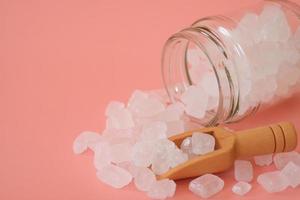 azúcar de roca de caramelo blanco o azúcar de cristal en cuchara de madera y frasco de vidrio aislado sobre fondo rosa. el azúcar de roca blanca se derrite y luego se deja cristalizar en una piedra preciosa semitranslúcida. foto