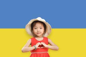 Little girl wear hat showing hands in heart shape on Ukraine flag color background. Stop the war for peace. Love Ukraine concept. photo