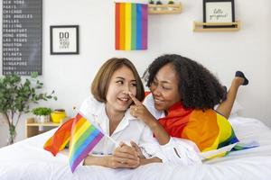 Couple of same sex marriage from difference races with the LGBTQ rainbow flag for pride month while lying down together on bed to promote equality and differences of homosexual and discrimination photo