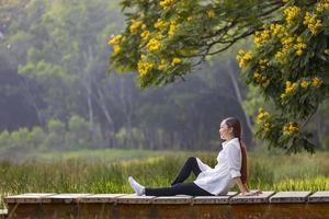 Asian woman sitting on the edge of dock with peaceful natural park during summer with yellow flower blossom for serene and relaxation outdoor recreation photo