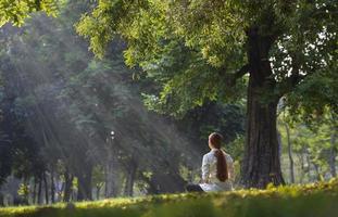 Back of woman relaxingly practicing meditation in the forest to attain happiness from inner peace wisdom with beam of sun light for healthy mind and soul photo