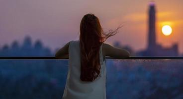 mujer mirando y disfrutando de la vista del atardecer desde el balcón con la puesta de sol detrás de un rascacielos en el ajetreado centro urbano con soledad por soledad, soledad y soñando con un estilo de vida libre foto
