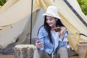 Asian woman is using mobile phone while drinking coffee at her tent while camping outdoor during summer time in national park for adventure and active travel concept