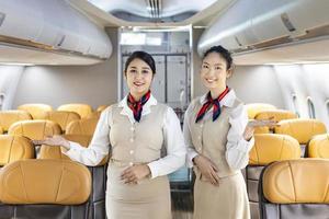equipo de asistente de vuelo asiático posando con una sonrisa en medio del pasillo dentro del avión para dar la bienvenida al pasajero a bordo con asiento en el fondo foto