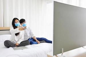 Couple wearing facial mask watching news on television while staying at home during quarantine from coronavius or covid-19 pandemic with copy space photo