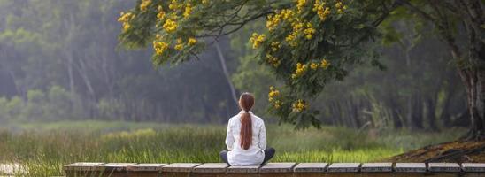 vista panorámica posterior de una mujer practicando relajantemente la meditación en el parque público para alcanzar la felicidad de la sabiduría de la paz interior con flores amarillas en verano foto