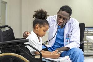 médico afroamericano está diagnosticando al niño discapacitado en silla de ruedas usando estetoscopio para chequeo pulmonar y efecto secundario sobre coronavirus foto