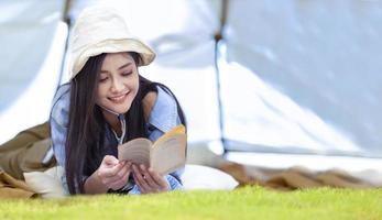 Asian woman is reading a book in her tent while camping outdoor during summer time in national park for adventure and active travel concept