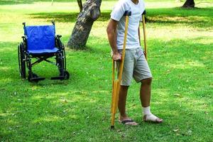 Los pacientes con lesiones en el tobillo se envuelven con un vendaje en el tobillo y usan muletas para caminar sobre el césped, que tiene una silla de ruedas detrás. concepto médico y de salud. foto