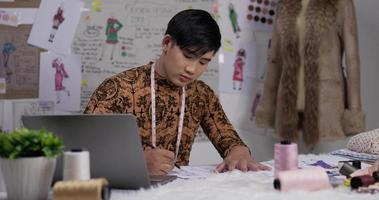 retrato de un diseñador de ropa asiático trabajando en una laptop y dibujando un boceto de ropa en el estudio. el pequeño empresario de inicio está en proceso de crear una nueva colección de ropa. video